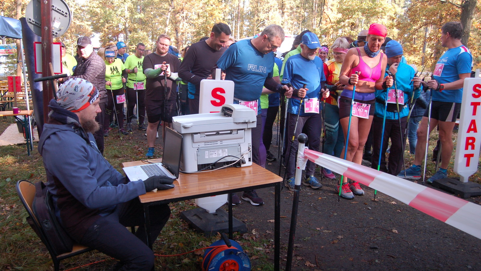 sędzia przy stoliku sędziowskim, w tle zawodnicy nordic walking ustawieni do startu